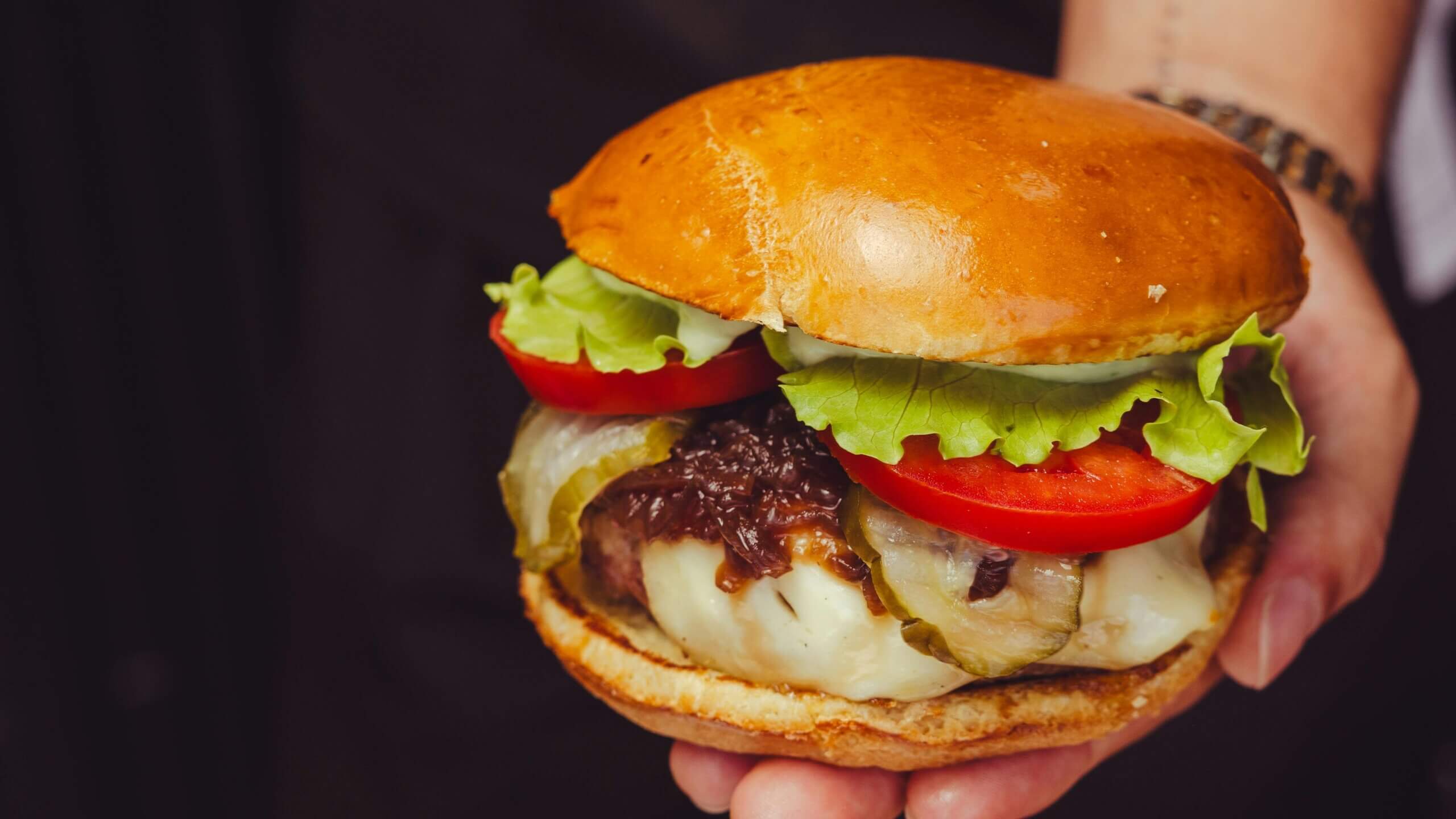 Tangan memegang burger keju dengan selada, tomat, acar, dan bawang karamel di atas roti brioche.