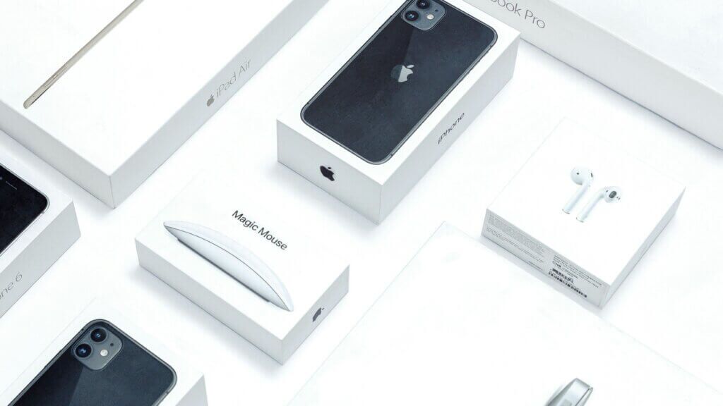Kotak produk Apple termasuk iPhone, iPad Air, Magic Mouse, AirPods, dan MacBook Pro pada permukaan putih