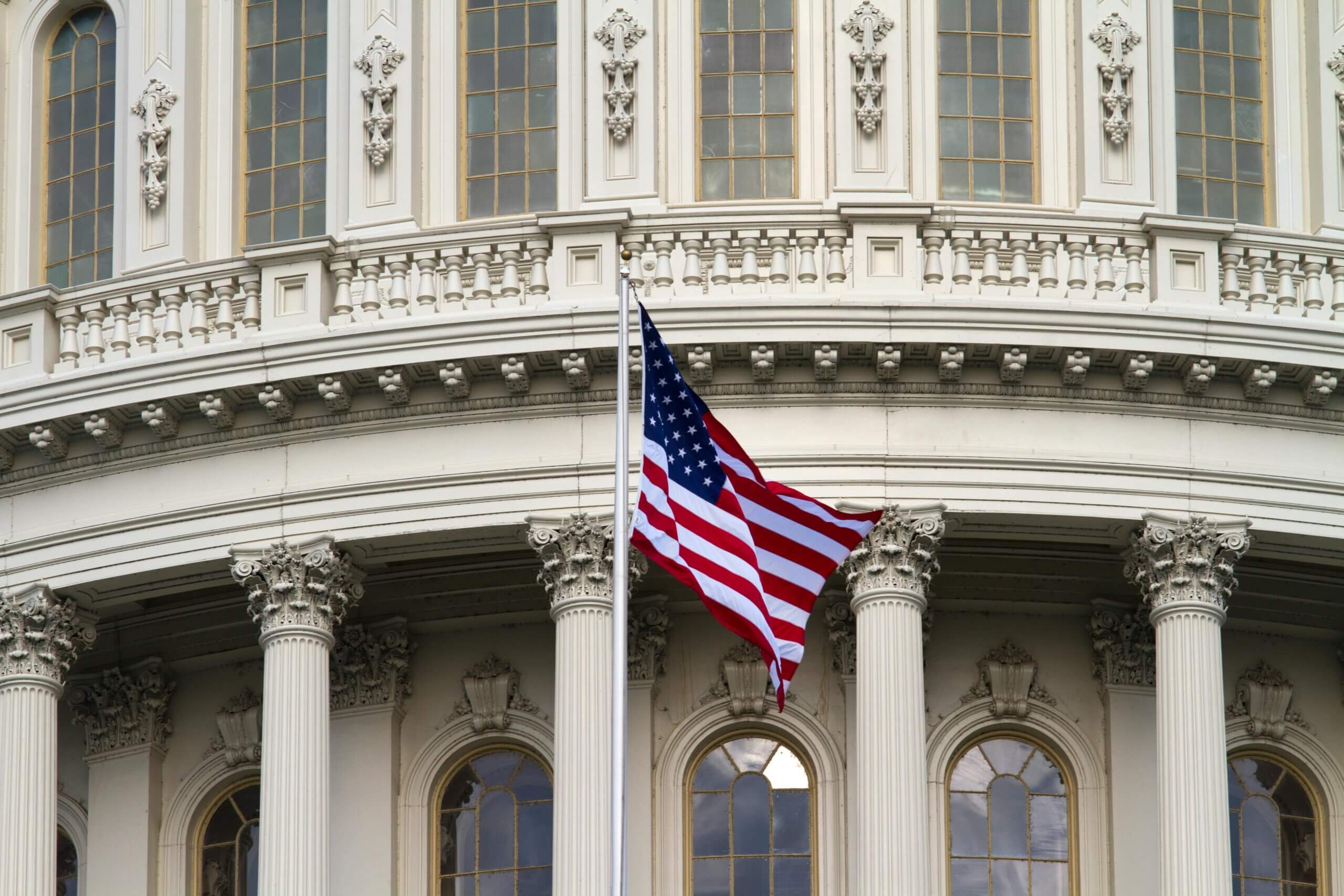 Bendera Amerika berkibar di depan gedung Capitol Amerika Serikat dengan tiang-tiang Korintus dan jendela melengkung