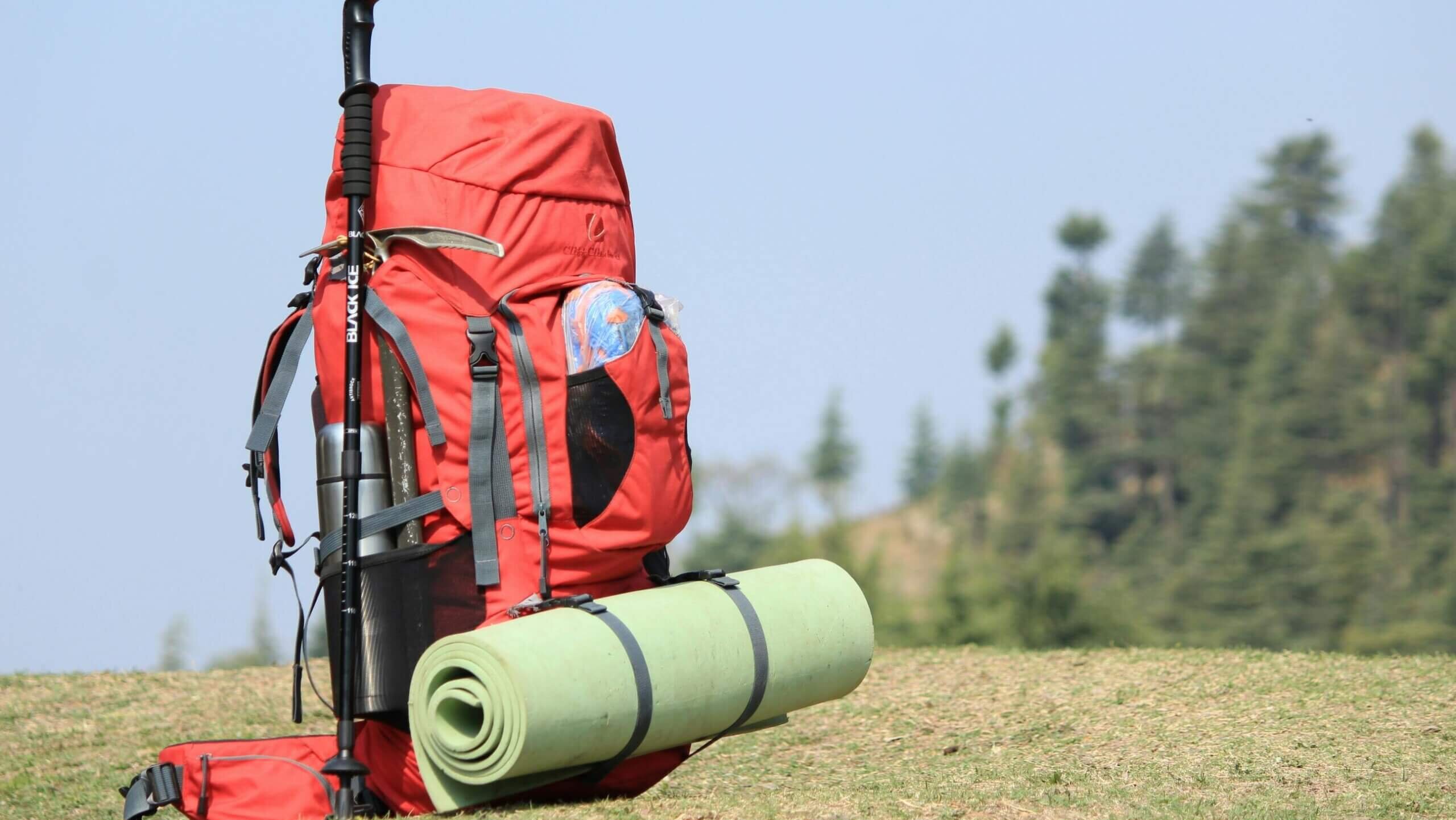 Ransel hiking berwarna merah dengan alas tidur hijau dan tiang trekking Black Ice di rumput dengan latar belakang pepohonan.