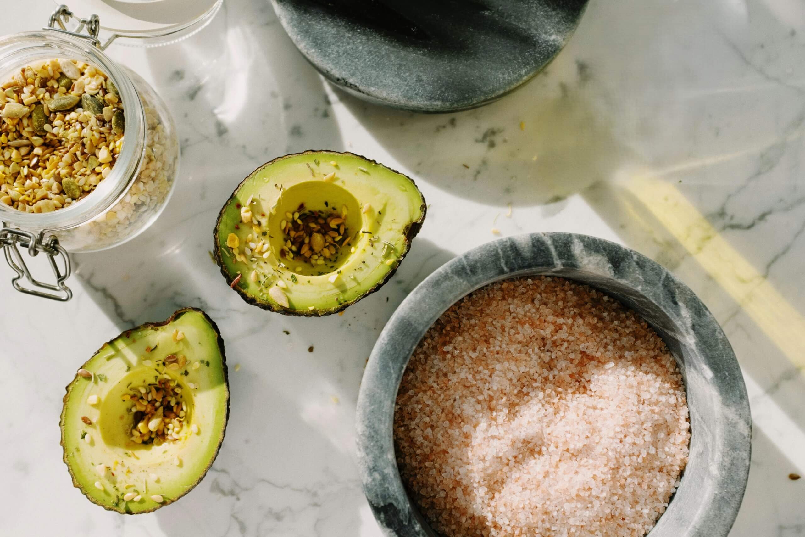 Dua bagian alpukat ditaburi biji-bijian dan rempah-rempah di samping toples berisi campuran biji-bijian dan semangkuk garam Himalaya merah muda di permukaan marmer