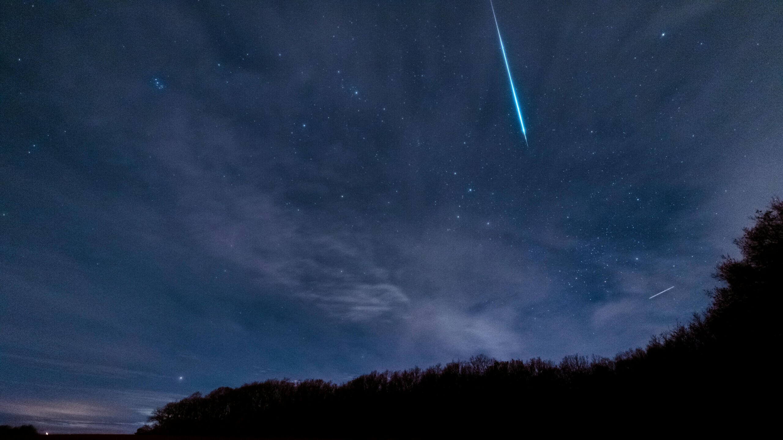 Meteor terang melesat melintasi langit malam berbintang di atas siluet hutan gelap.