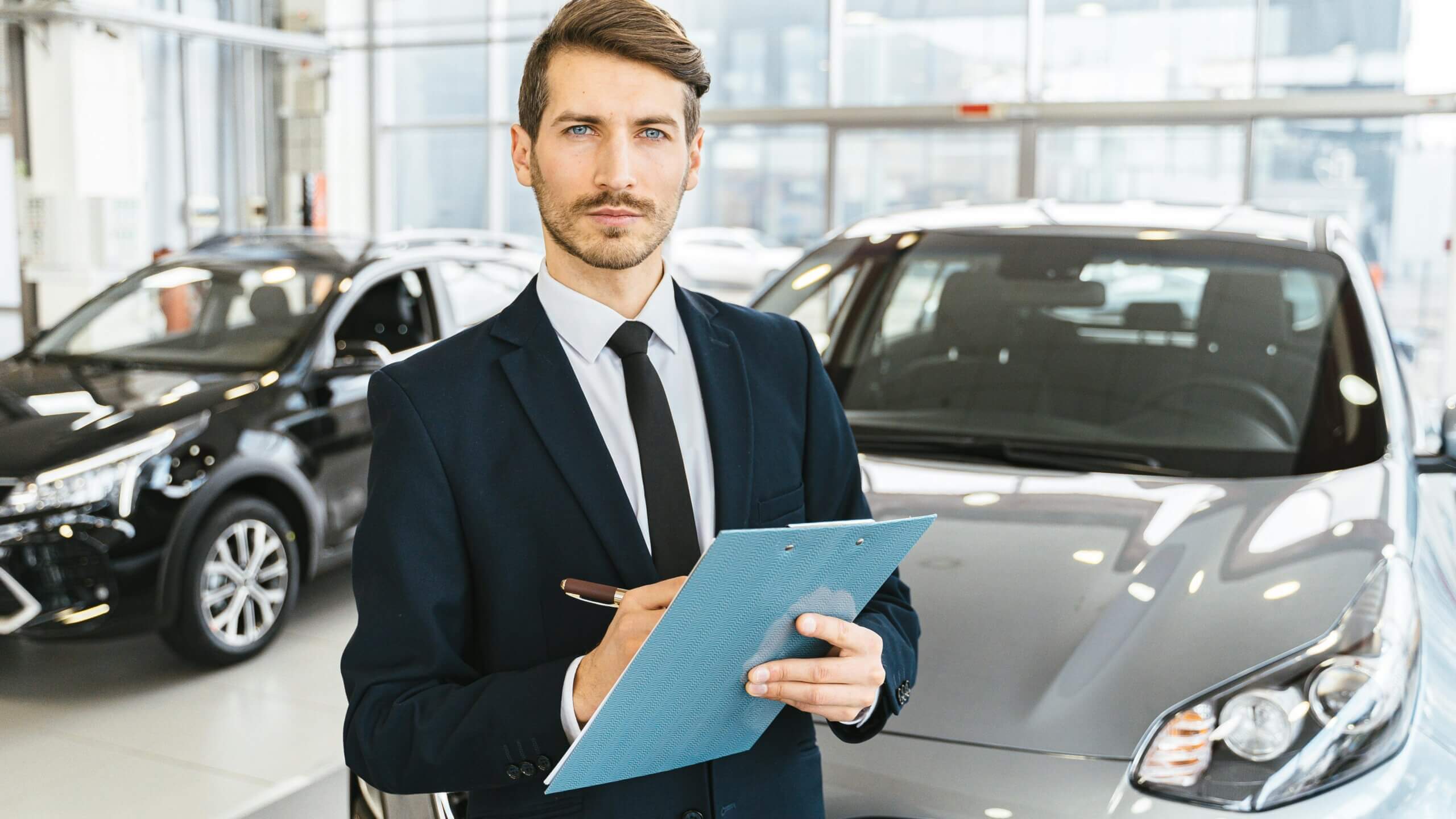 Pria berjas memegang papan klip berdiri di ruang pamer dealer mobil dengan mobil di latar belakang