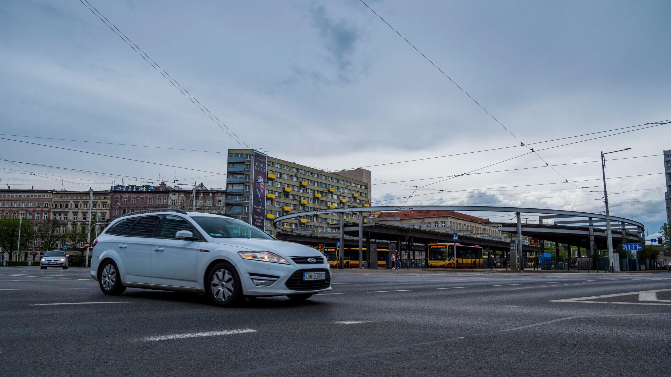 Station wagon White Ford melaju di jalan kota dekat stasiun bus dengan kabel trem di atas kepala dan gedung apartemen.