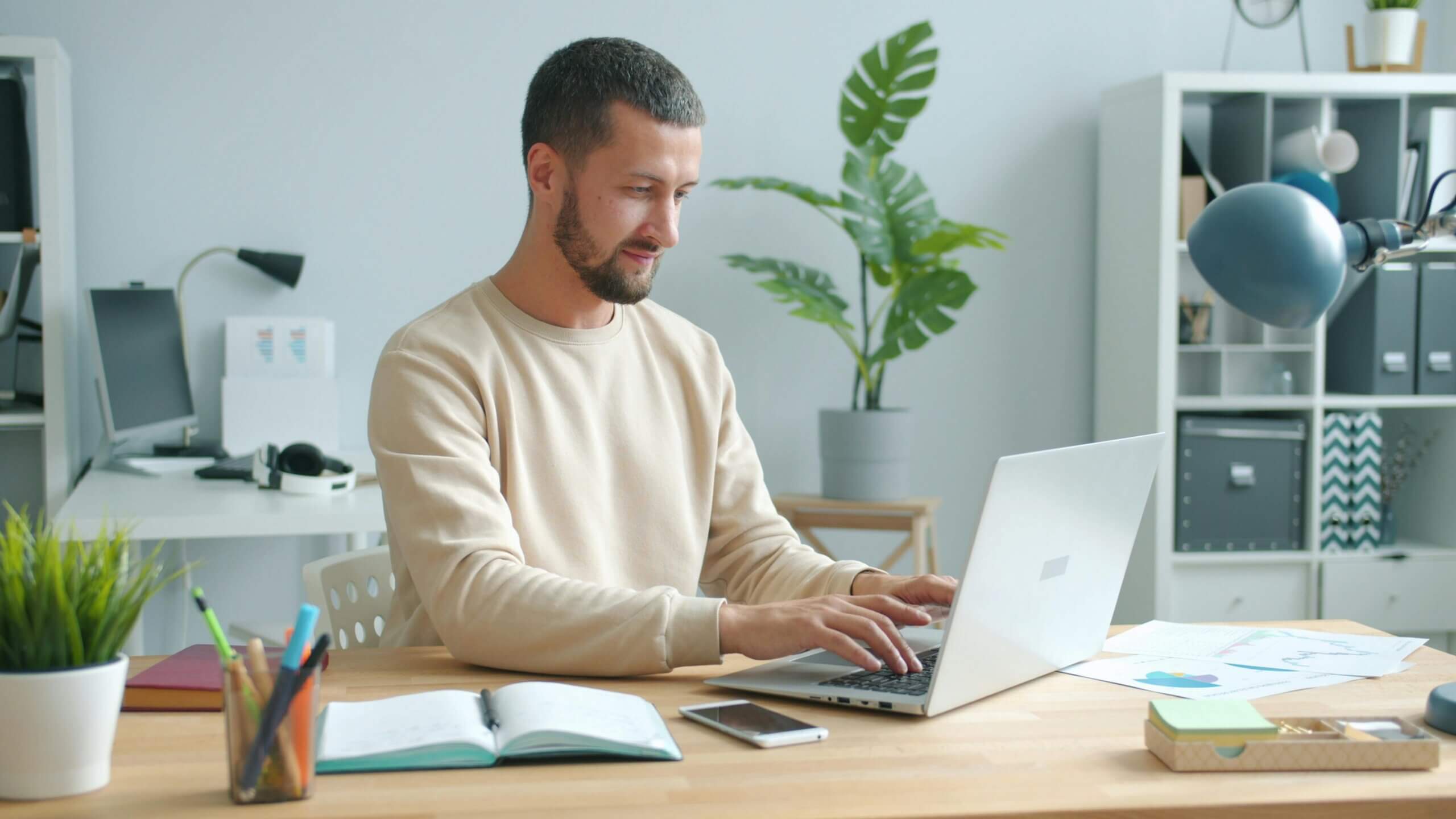 Pria berjaket krem ​​mengetik di laptop di meja dengan notebook, ponsel pintar, dan perlengkapan kantor di ruang kerja modern
