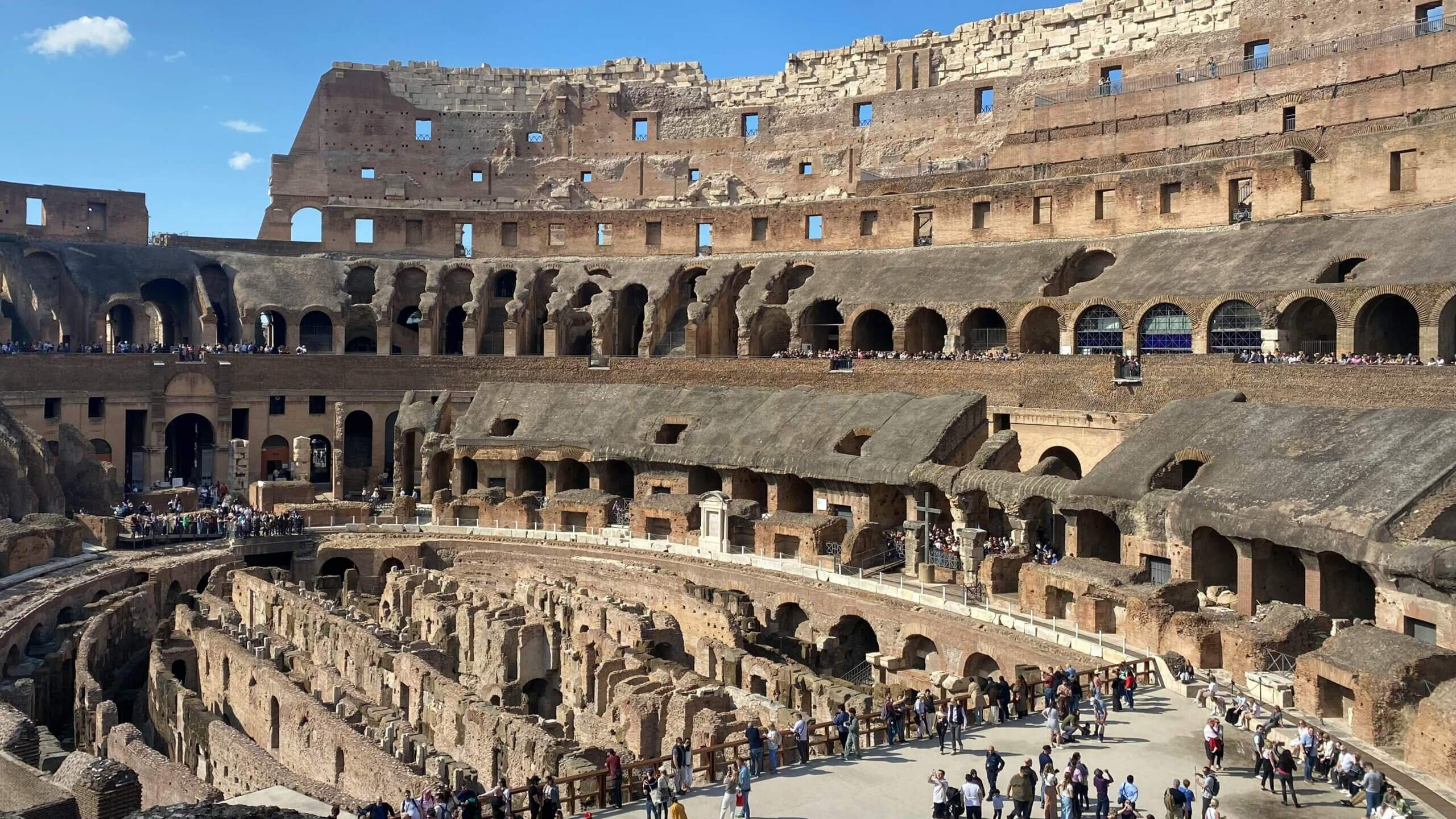 Pemandangan interior Colosseum di Roma dengan wisatawan menjelajahi amfiteater kuno di bawah langit biru cerah.