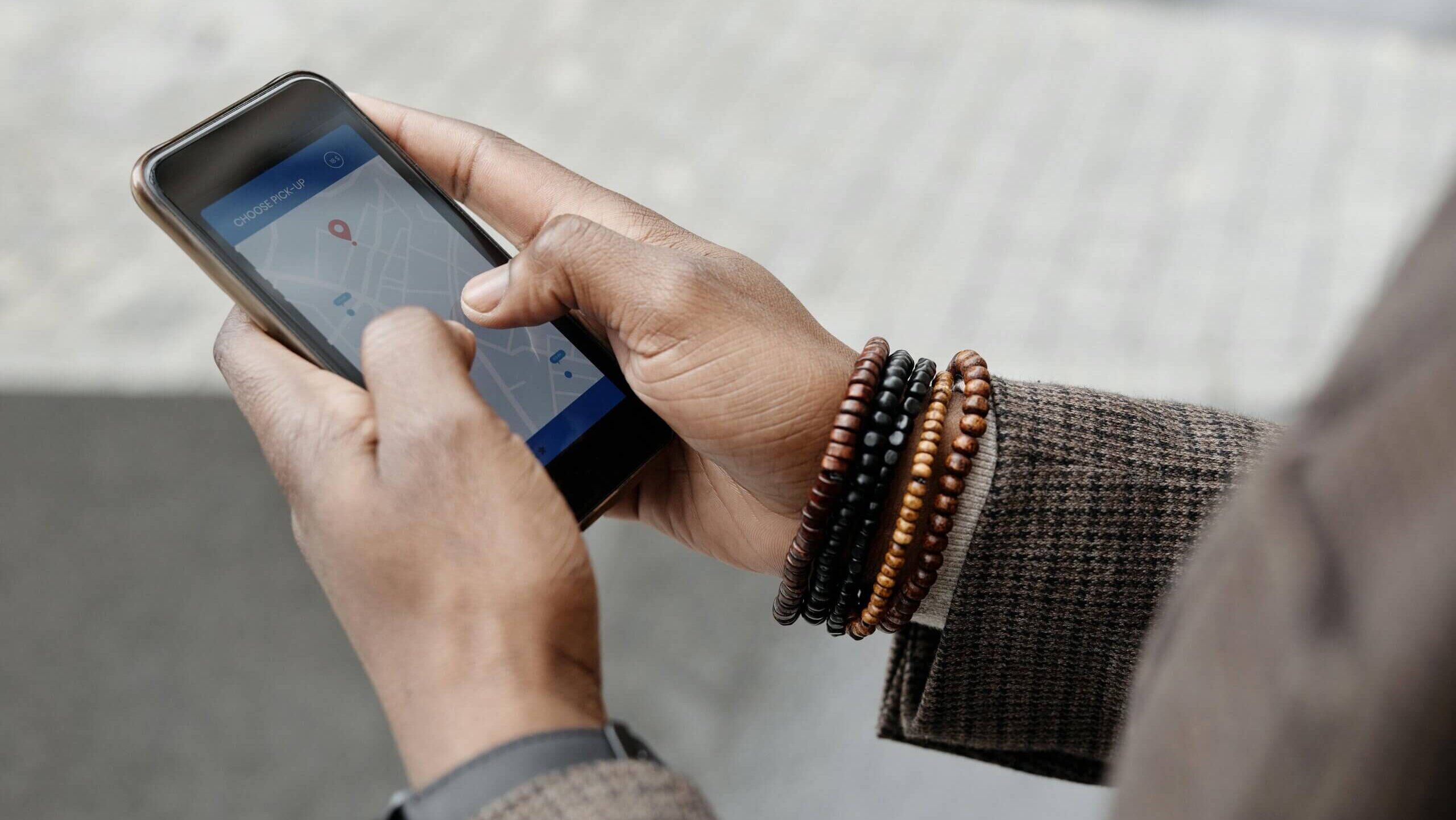 Orang yang menggunakan ponsel cerdas dengan aplikasi peta terbuka, memilih lokasi penjemputan, mengenakan gelang manik-manik dan jaket kotak-kotak.