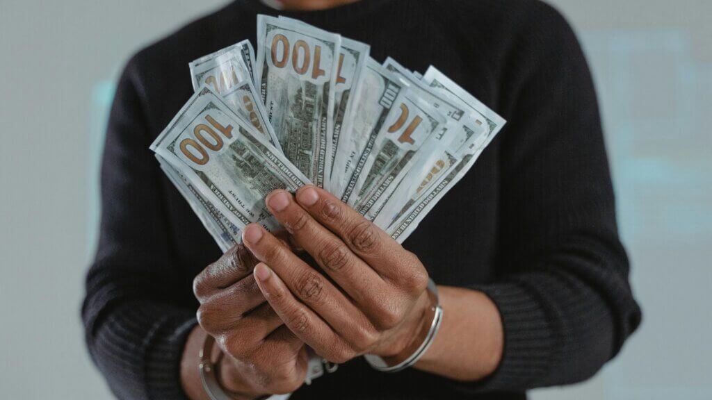 Orang yang memegang kipas uang kertas 100 dolar AS mengenakan sweter hitam dan gelang perak
