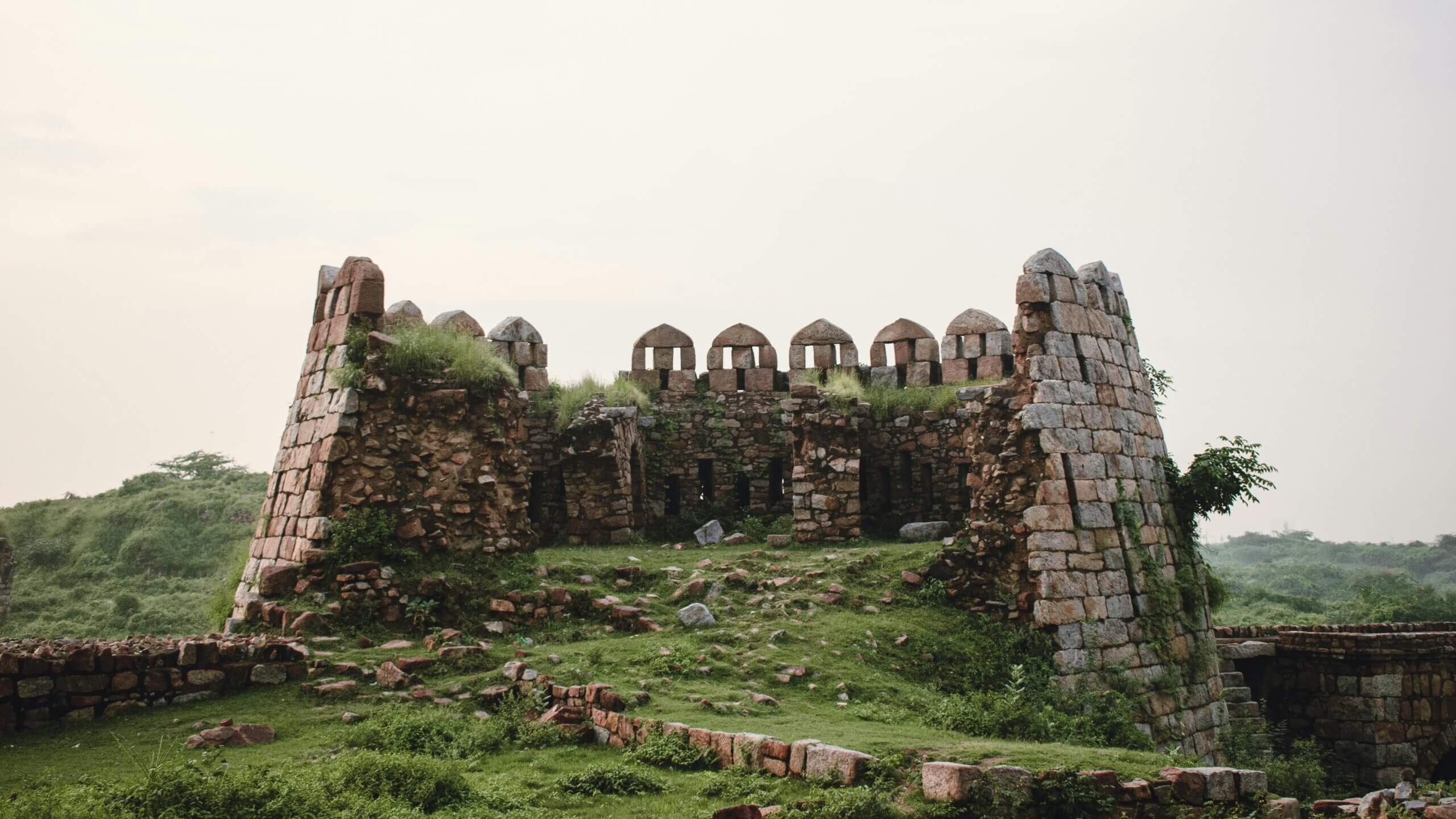 Dinding benteng batu yang hancur dengan benteng melengkung dikelilingi oleh rumput hijau dan tumbuh-tumbuhan.