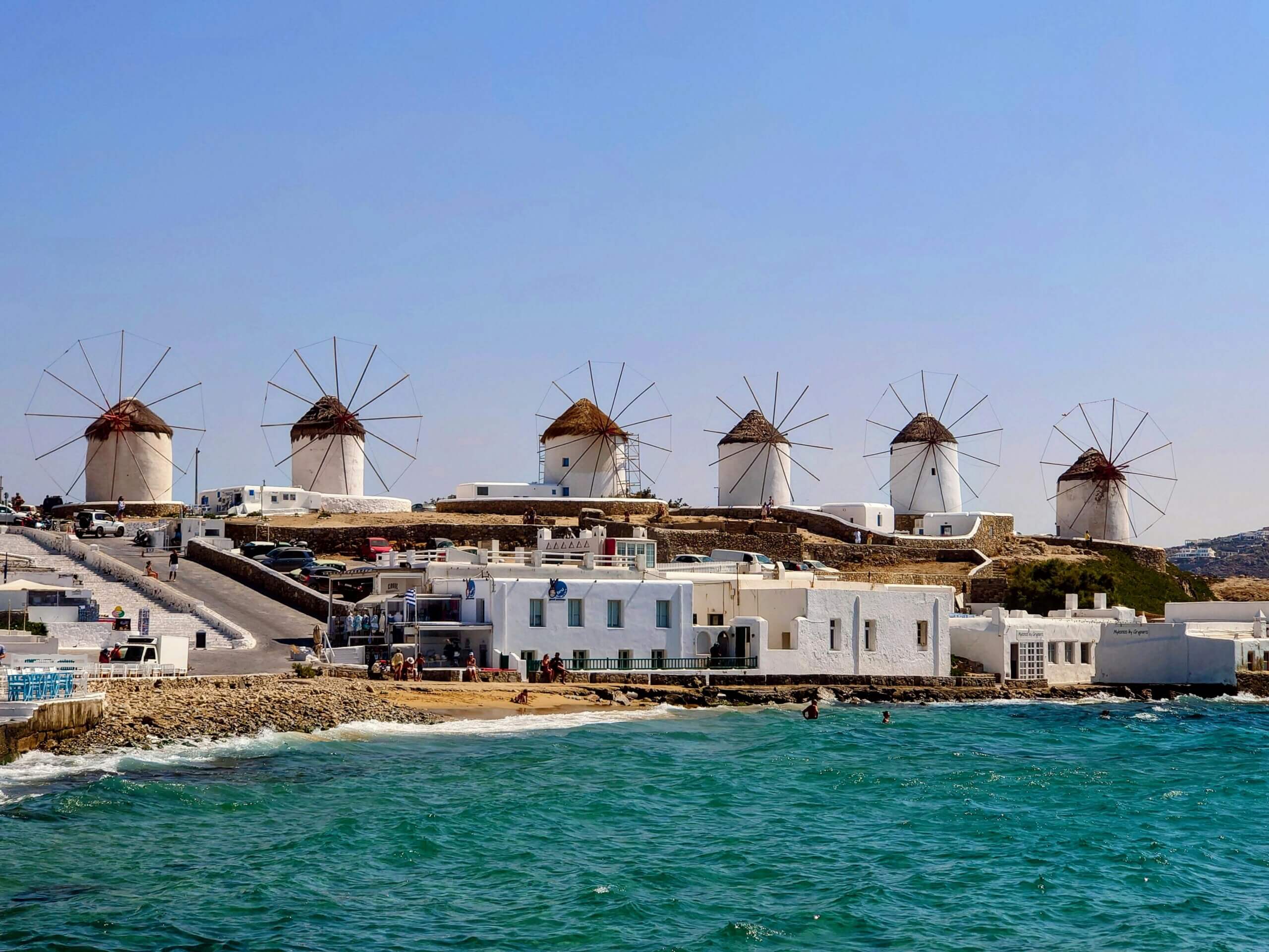 Enam kincir angin putih tradisional dengan atap jerami di sebuah bukit yang menghadap ke laut di Mykonos, Yunani.