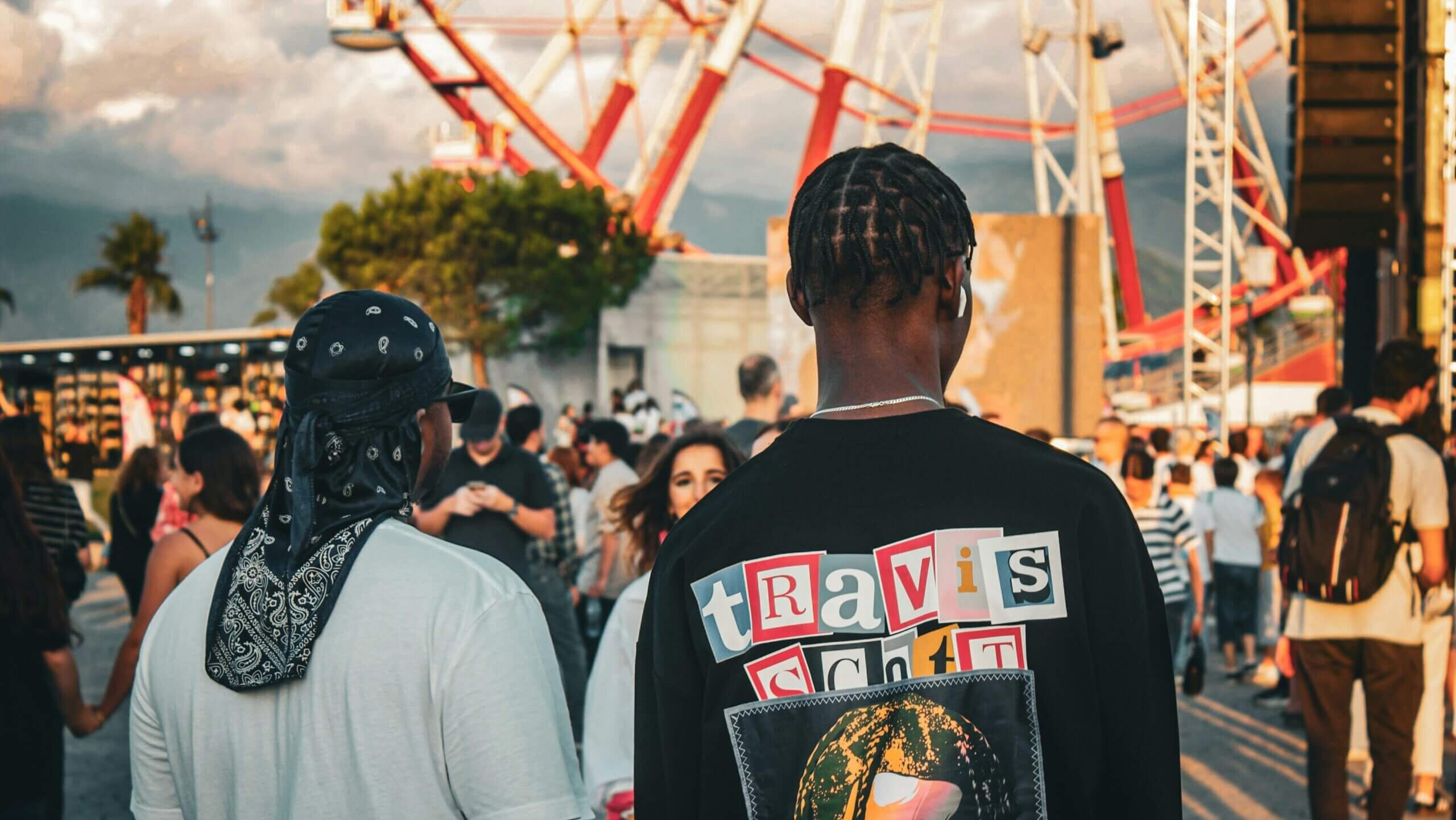 Dua pria berjalan di sebuah acara outdoor, salah satunya mengenakan kemeja hitam Travis Scott dengan potret di bagian belakang.