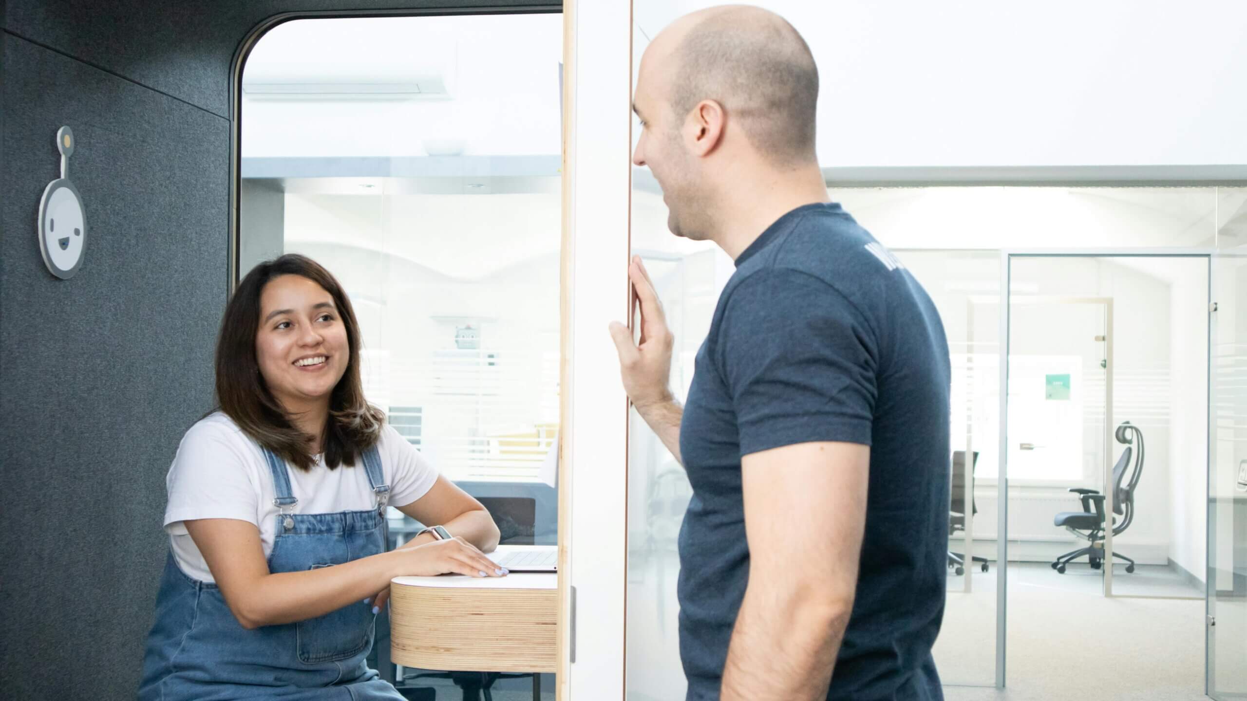Wanita berbaju denim tersenyum pada pria botak dengan kemeja biru tua saat berbincang di kantor modern.