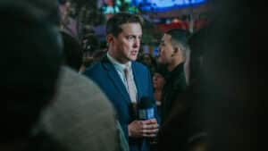 News reporter in a blue suit holding a 9 News microphone speaking to a man in a crowded outdoor setting.