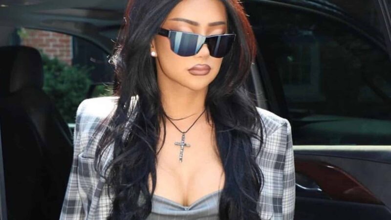 Woman with long black hair wearing large black sunglasses, a plaid blazer, and a cross necklace stepping out of a car