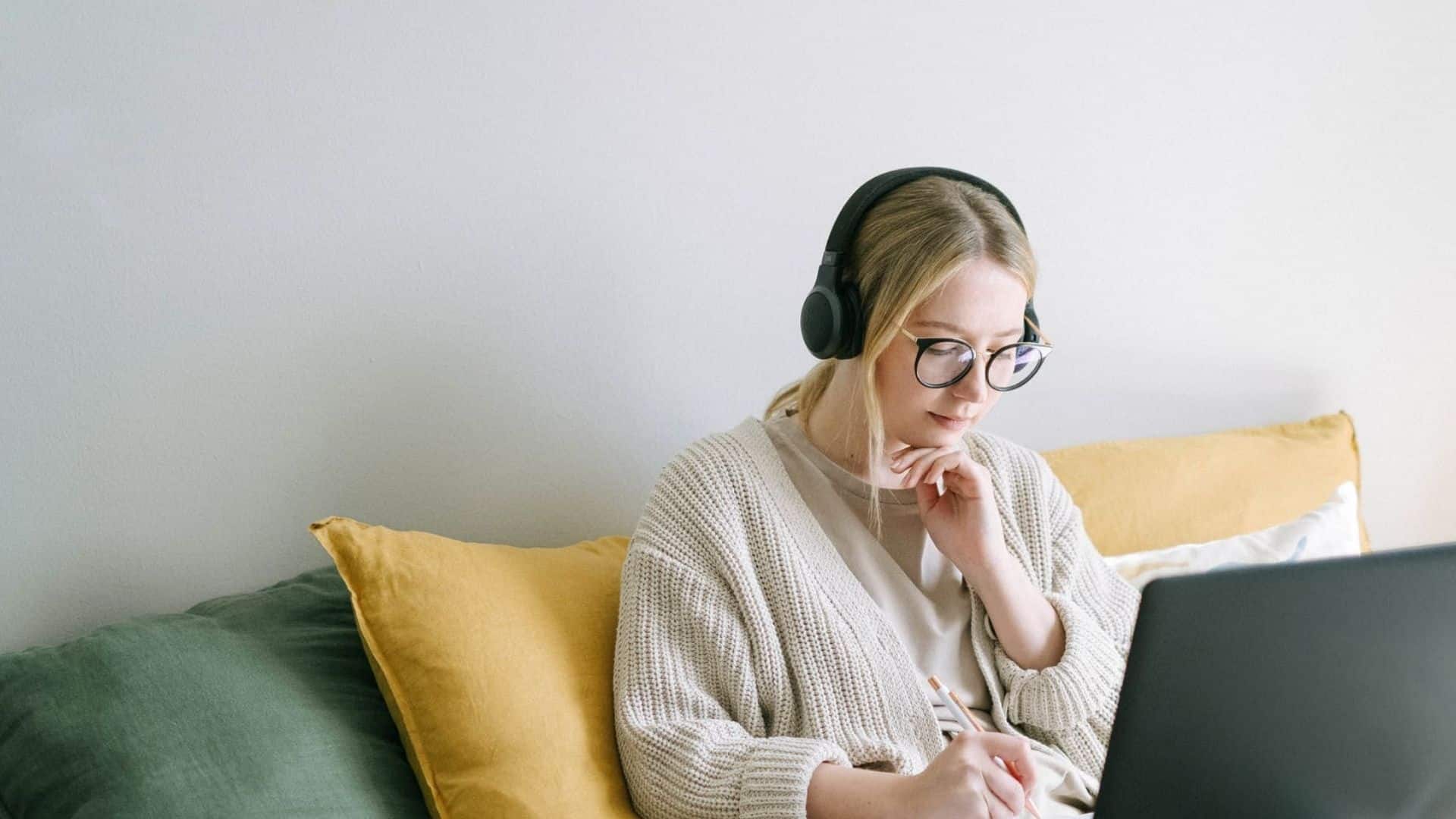 Young woman wearing glasses and headphones, writing notes while using a laptop on a couch with yellow and green pillows.