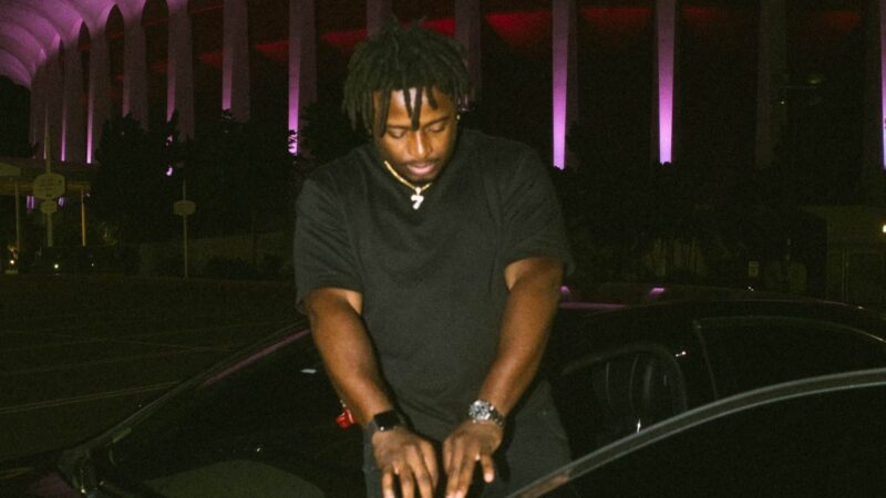Man with dreadlocks wearing black shirt and gold necklace standing by a car at night with purple-lit columns in the background