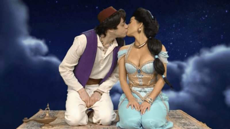 Two people dressed as Aladdin and Jasmine kissing on a flying carpet with a starry night sky background.