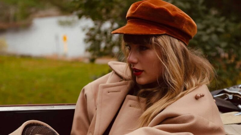Taylor Swift wearing a brown cap and beige coat sitting outdoors near a body of water.