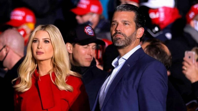 Ivanka Trump in red coat and Donald Trump Jr. in navy suit at a political rally with red "Make America Great Again" hats in background.