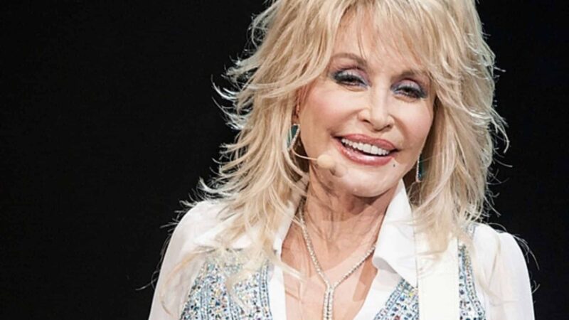 Dolly Parton smiling on stage wearing a white shirt and sparkly vest with a microphone headset.