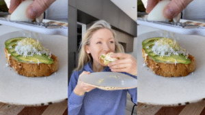 Woman in blue sweater eating avocado toast topped with grated cheese in a modern kitchen