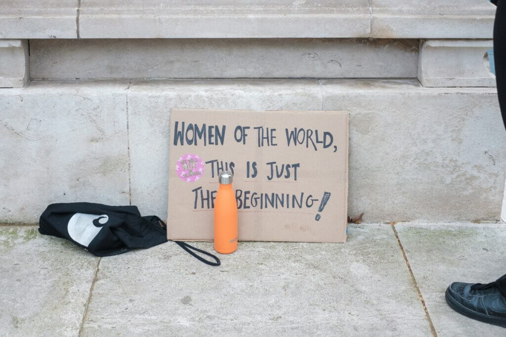 Cardboard sign reading "Women of the world, this is just the beginning!" with a pink sticker and an orange water bottle nearby.