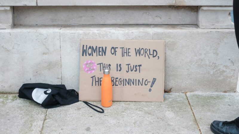 Cardboard sign reading "Women of the world, this is just the beginning!" with a pink sticker and an orange water bottle nearby.