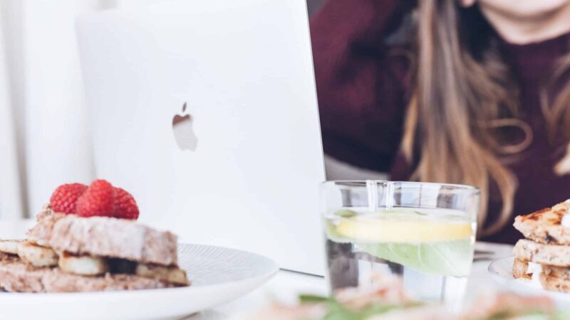 Apple laptop on a table with plates of sandwiches and a glass of lemon water, a person in the background
