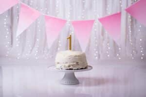 White frosted cake with a gold number 1 candle on a stand, pink gradient pennant banner and fairy lights background