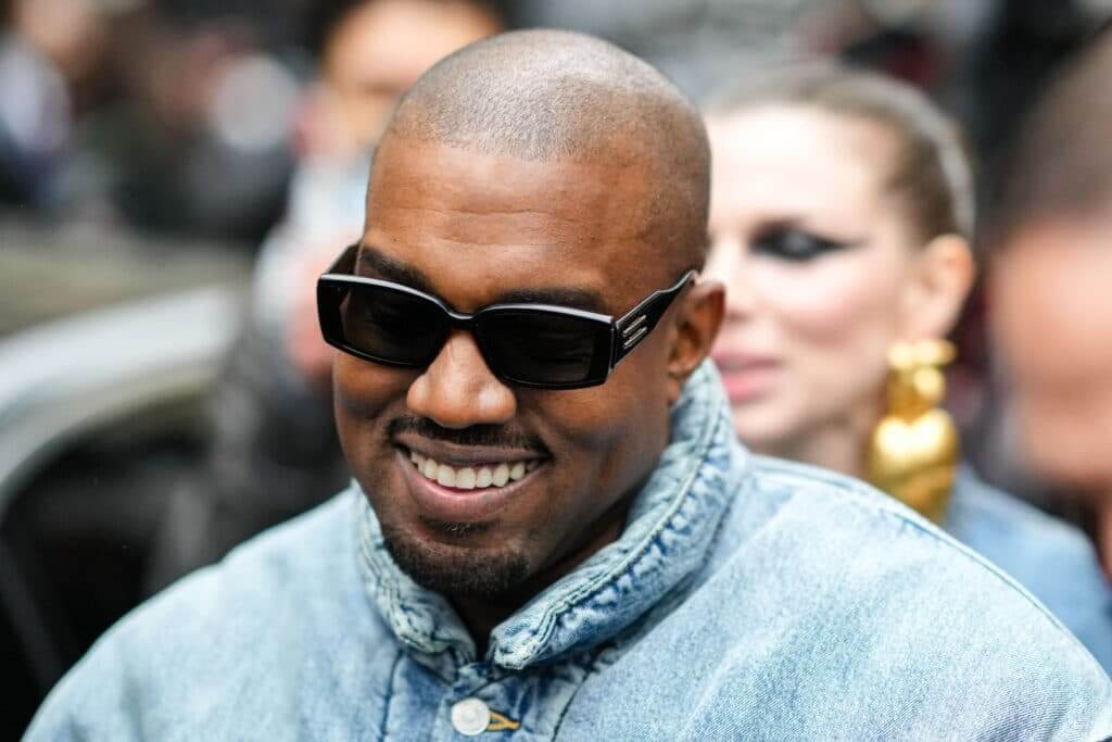 Kanye West smiling in black sunglasses and a light denim jacket with a blurred woman in the background.