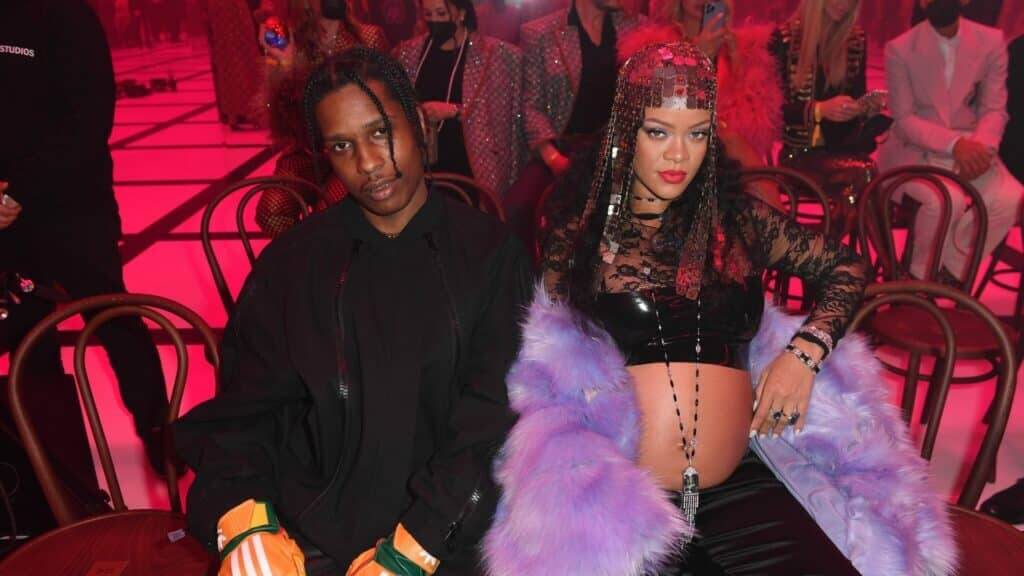 A$AP Rocky and pregnant Rihanna seated at a fashion event with red lighting and stylish outfits.