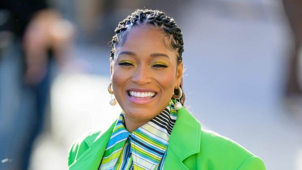 Keke Palmer smiling with braided hair, yellow eyeshadow, pearl hoop earrings, and a green jacket over a striped shirt
