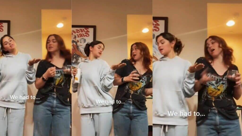 Two women dancing indoors, one in a light gray hoodie and the other in a black graphic t-shirt holding a drink.