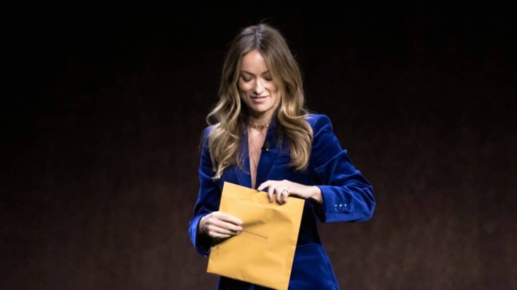 Woman in a blue velvet blazer opening a yellow envelope against a dark background