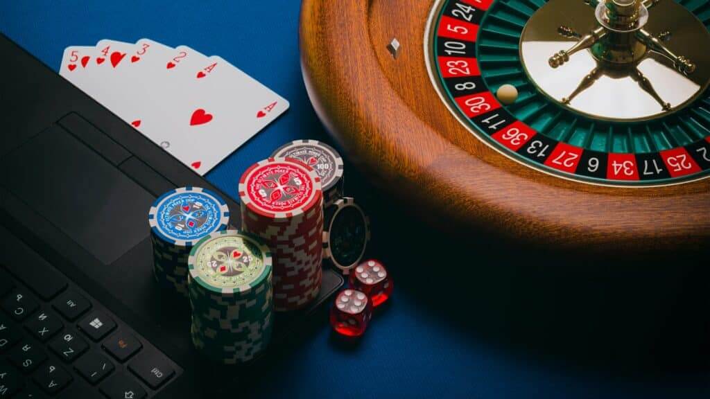 Roulette wheel, poker chips, red dice, playing cards, and laptop keyboard on blue surface.
