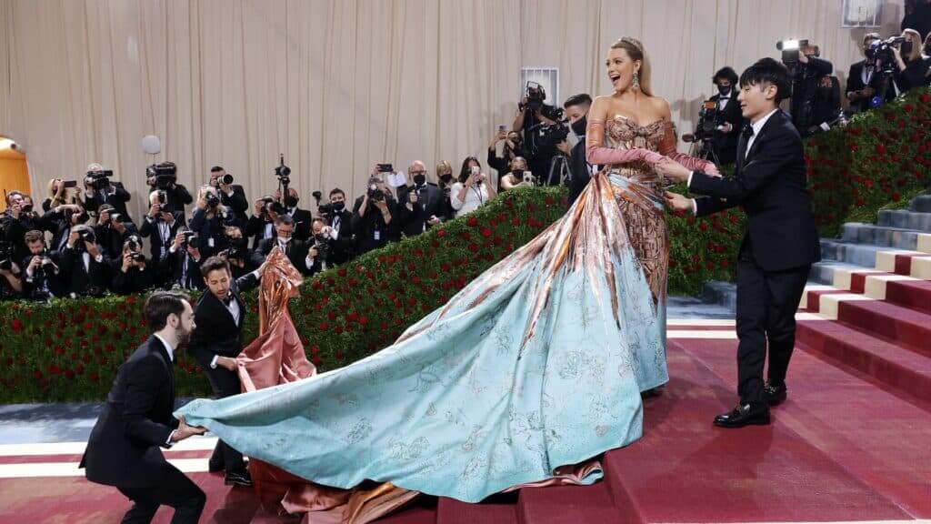 Blake Lively in a metallic and blue gown with a long train held by assistants on a red carpet with photographers in the background