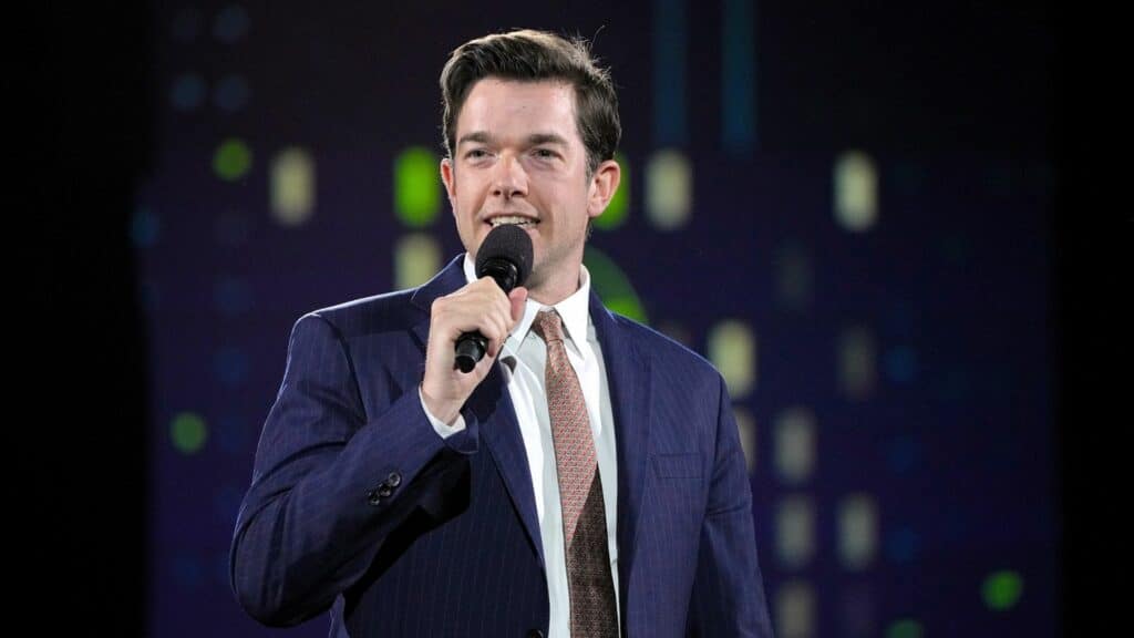 John Mulaney speaking into a microphone wearing a navy suit and patterned tie on stage