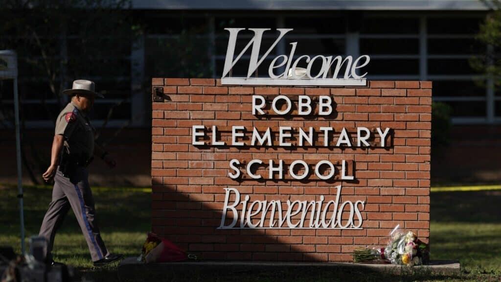 Texas state trooper walking past Robb Elementary School sign with flowers placed at its base