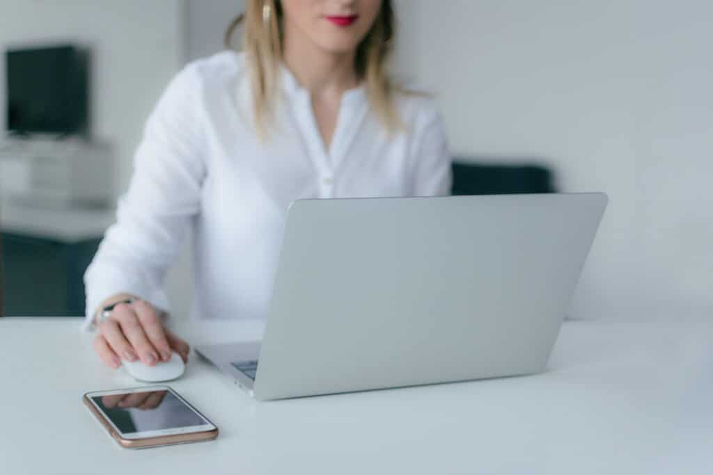 Woman in white shirt using a silver laptop with a smartphone on the desk and holding a white mouse