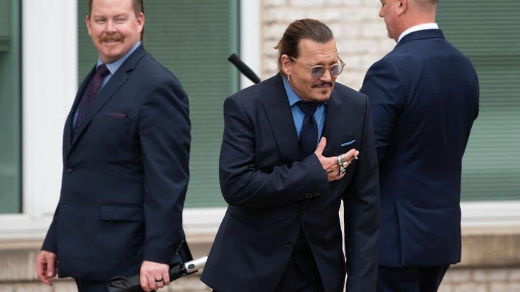 Johnny Depp in a navy suit and sunglasses, surrounded by two men in suits, outdoors near a building with green shutters.