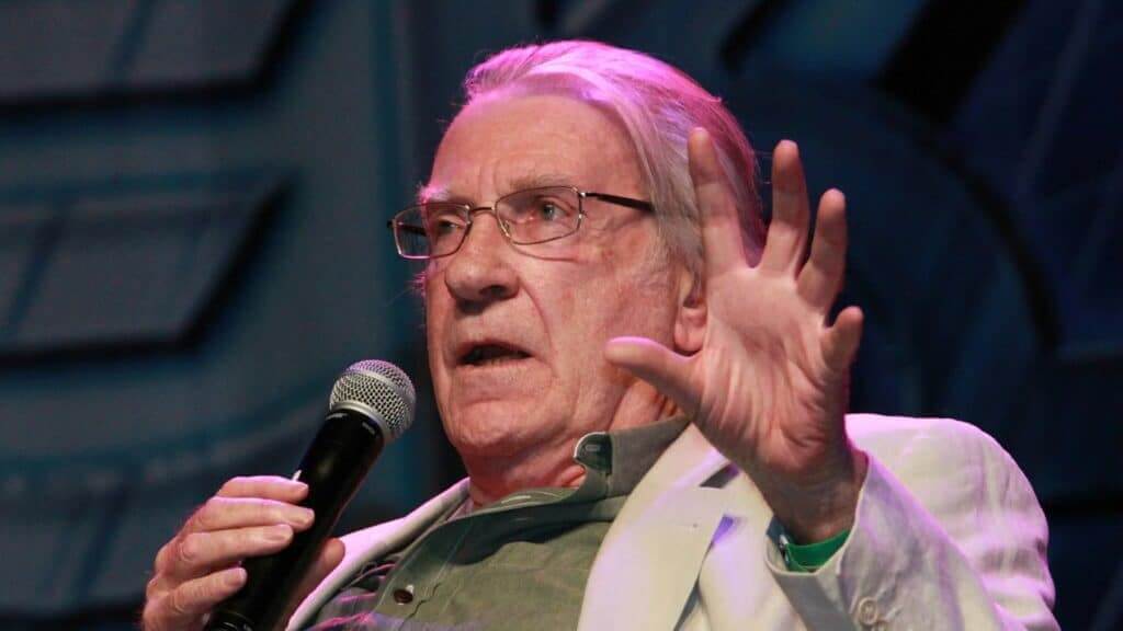 Elderly man with glasses speaking into a microphone and gesturing with his hand on stage.