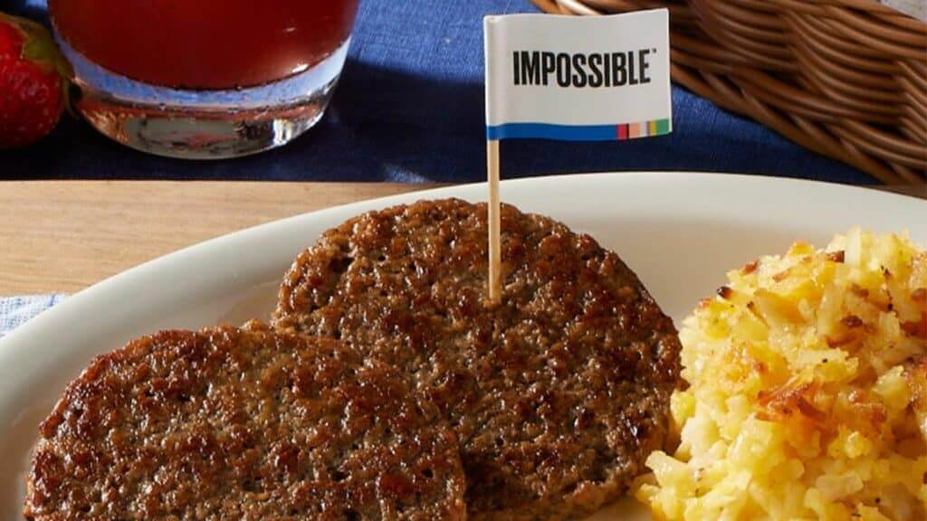 Two cooked Impossible burger patties with a flag on a plate next to hash browns and a glass of juice