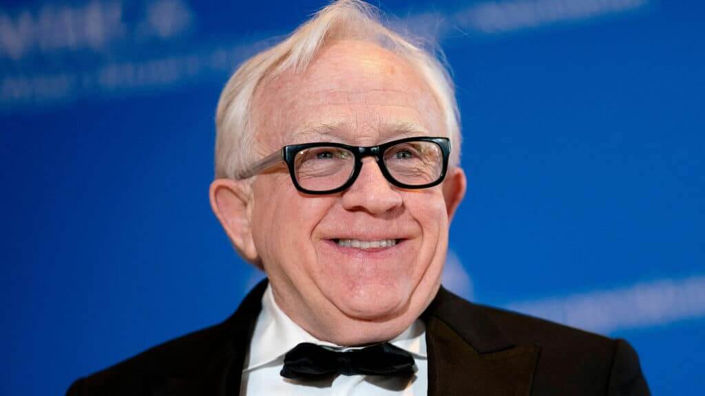 Smiling elderly man with white hair and black glasses wearing a tuxedo and bow tie against a blue background