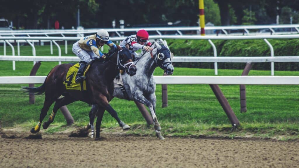 Two jockeys racing horses on a dirt track, one horse gray with a red helmet and the other dark with a yellow number 4 saddlecloth.
