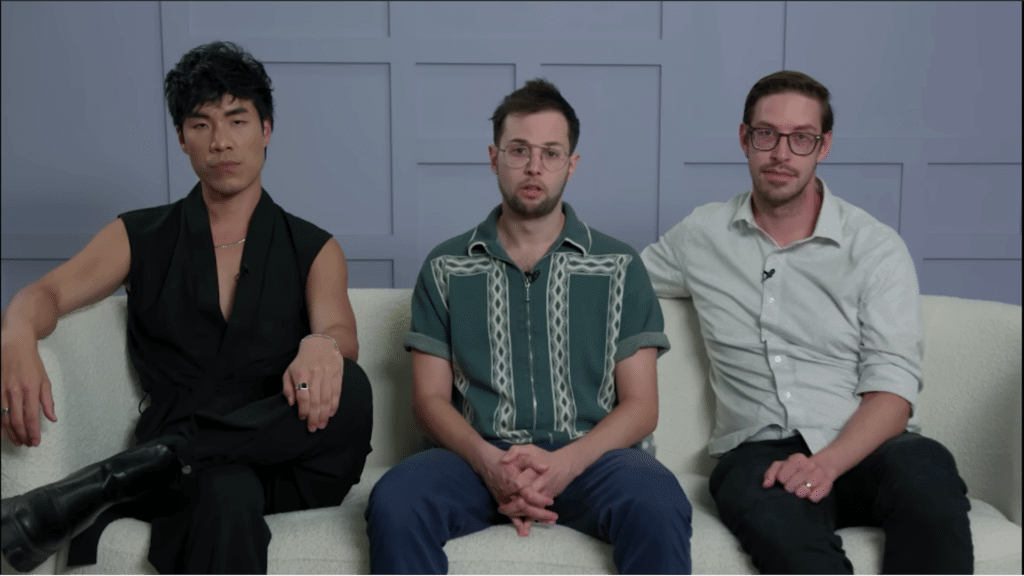 Three men sitting on a white couch against a gray paneled wall, one wearing a sleeveless black outfit, one in a green patterned shirt, and one in a light gray shirt.