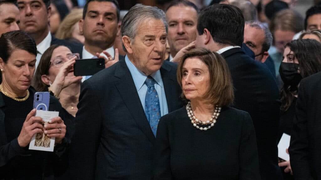 Nancy Pelosi and Paul Pelosi standing among a crowd, some people taking photos with phones.