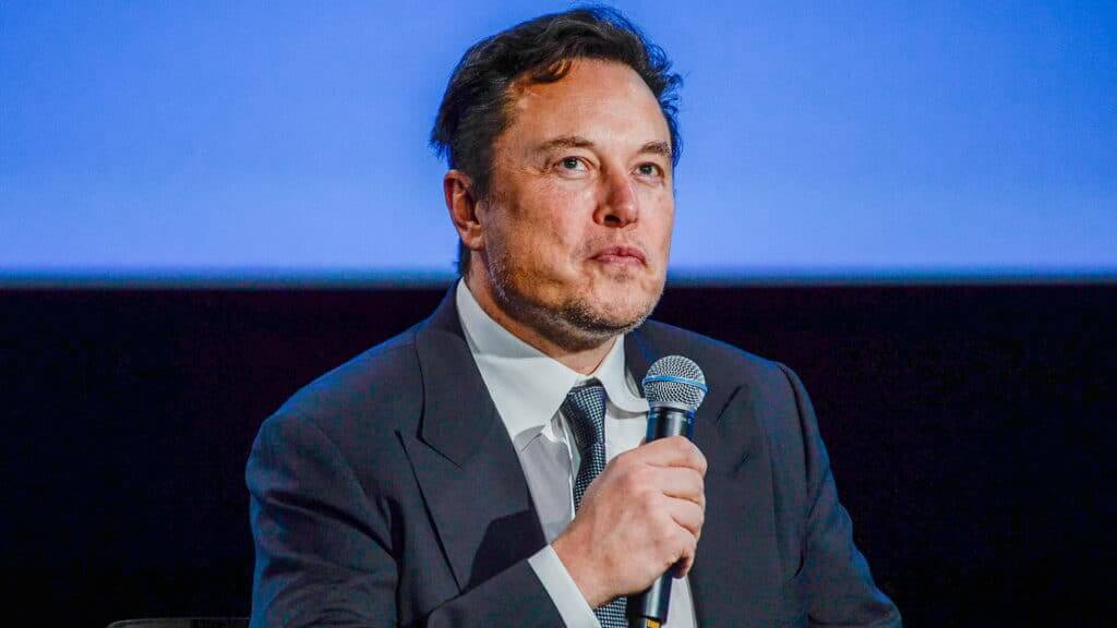 Elon Musk speaking into a microphone wearing a dark suit and tie against a blue background
