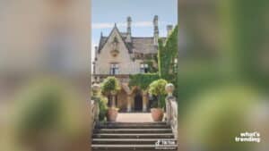 Stone mansion with ivy-covered walls and potted trees flanking a staircase entrance under a blue sky