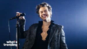 Harry Styles smiling on stage holding a microphone wearing a sparkly jacket and cross necklace