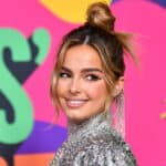 Addison Rae smiling in a silver sequined outfit with a top bun hairstyle against a colorful background